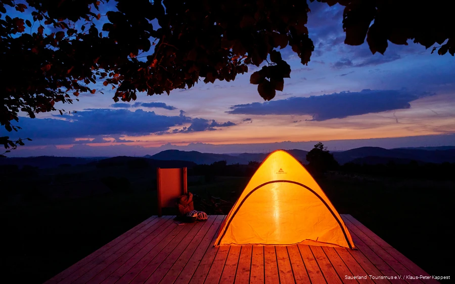 Ein von innen beleuchtetes Zelt steht auf einer Trekkingplattform, im Hintergrund ist eine Bergkette zu erkennen.