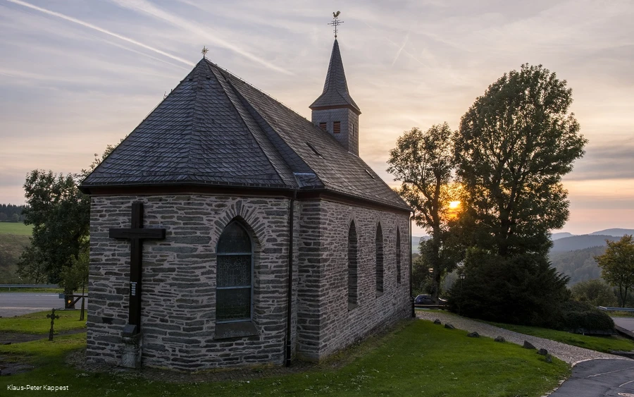 Eine steinerne Kapelle umgeben von grüner Wiese mit der untergehenden Sonne im Hintergrund.