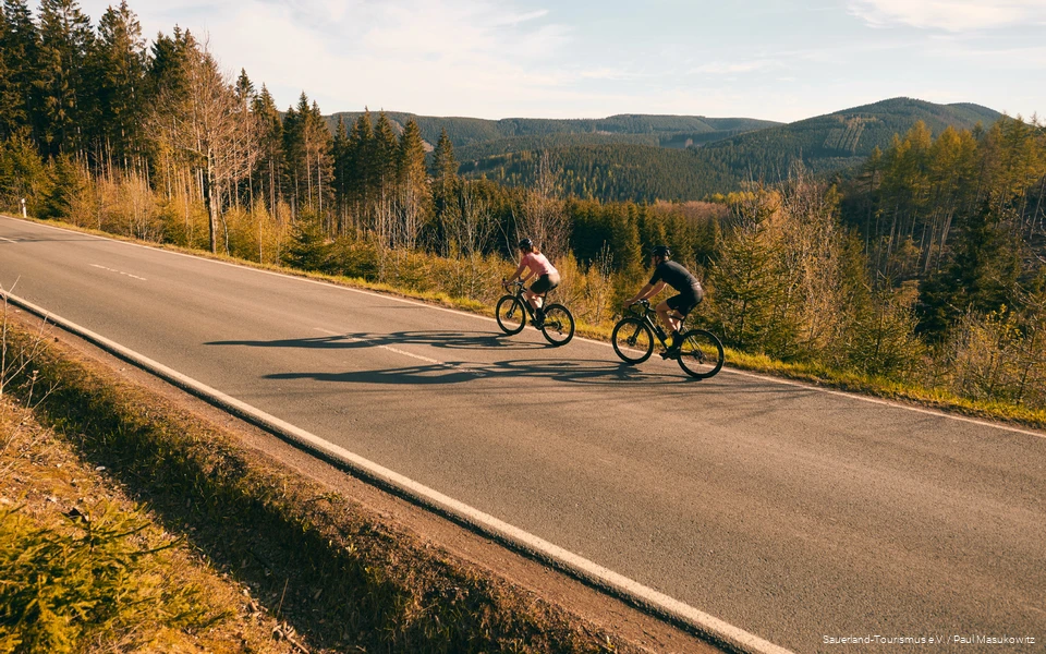 2 Rennradfahrende fahren eine Straße hinab, im Hintergrund ist die bergige Landschaft des Sauerlands zu sehen.