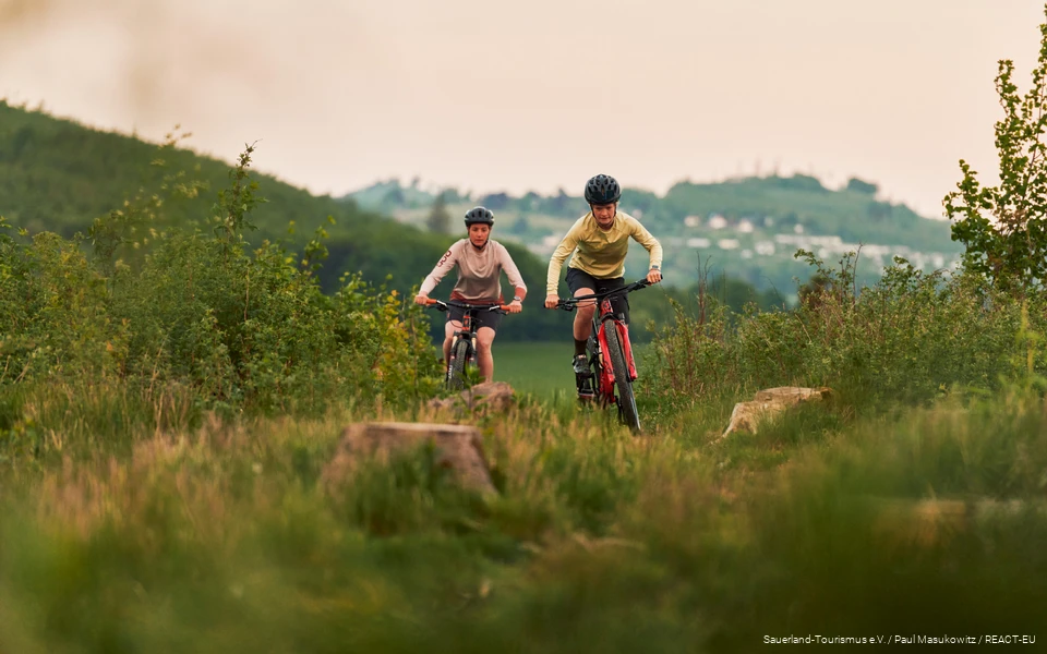 2 Mountainbikerinnen kommen einen Berg hoch, im Hintergrund sind Berge verschwommen zu sehen.